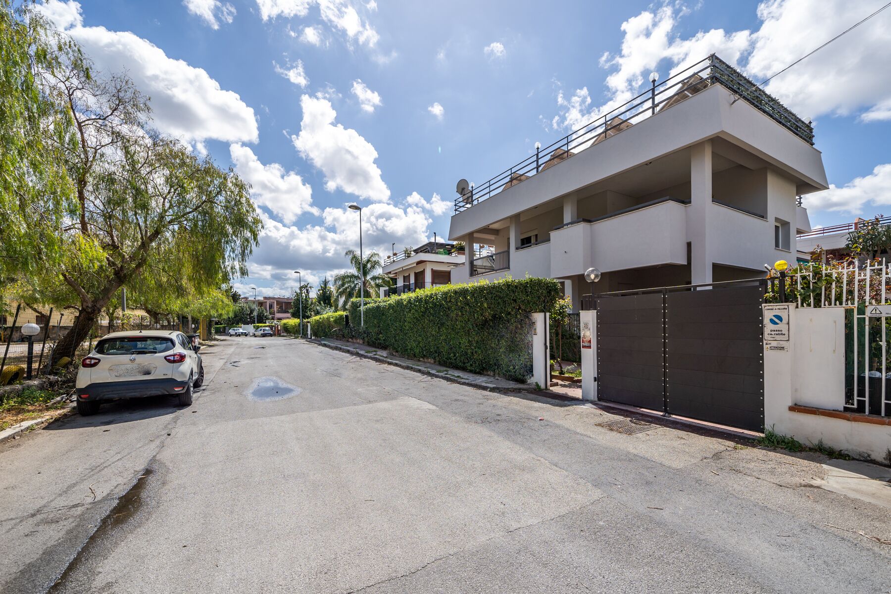 Une rue résidentielle avec un immeuble blanc, une entrée fermée par un portail et une voiture garée.