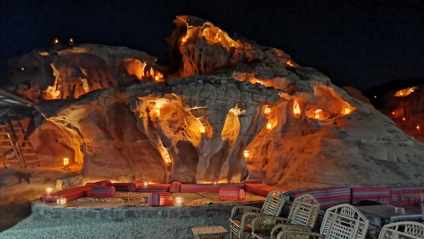 Formación rocosa con luces integradas, zona de estar con sillas y mesas, y una escalera apoyada en la roca.
