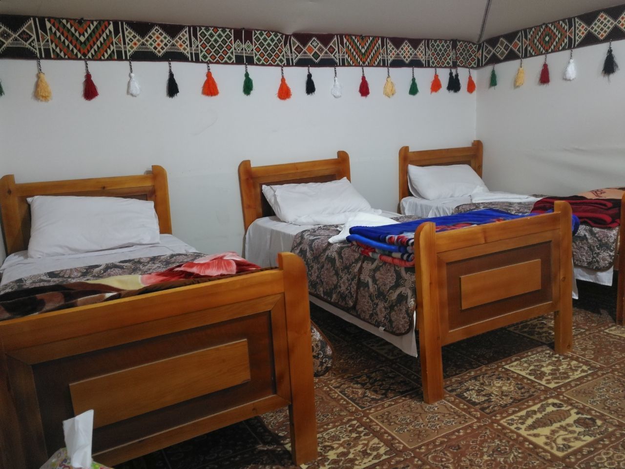 Une chambre avec trois lits en bois, chacun agrémenté d’un oreiller blanc et d’une couvre-lit à motifs. La chambre est dotée d’un tapis à motifs sur le sol et d’une décoration murale ornée de glands.