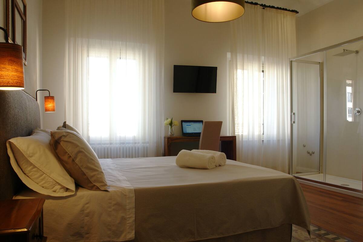 A hotel room with a bed, a desk with a computer, a TV, and a shower area.