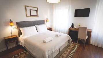 A neatly made bed with two pillows, a bedside table with a lamp, a desk with a chair, and a TV on the wall.