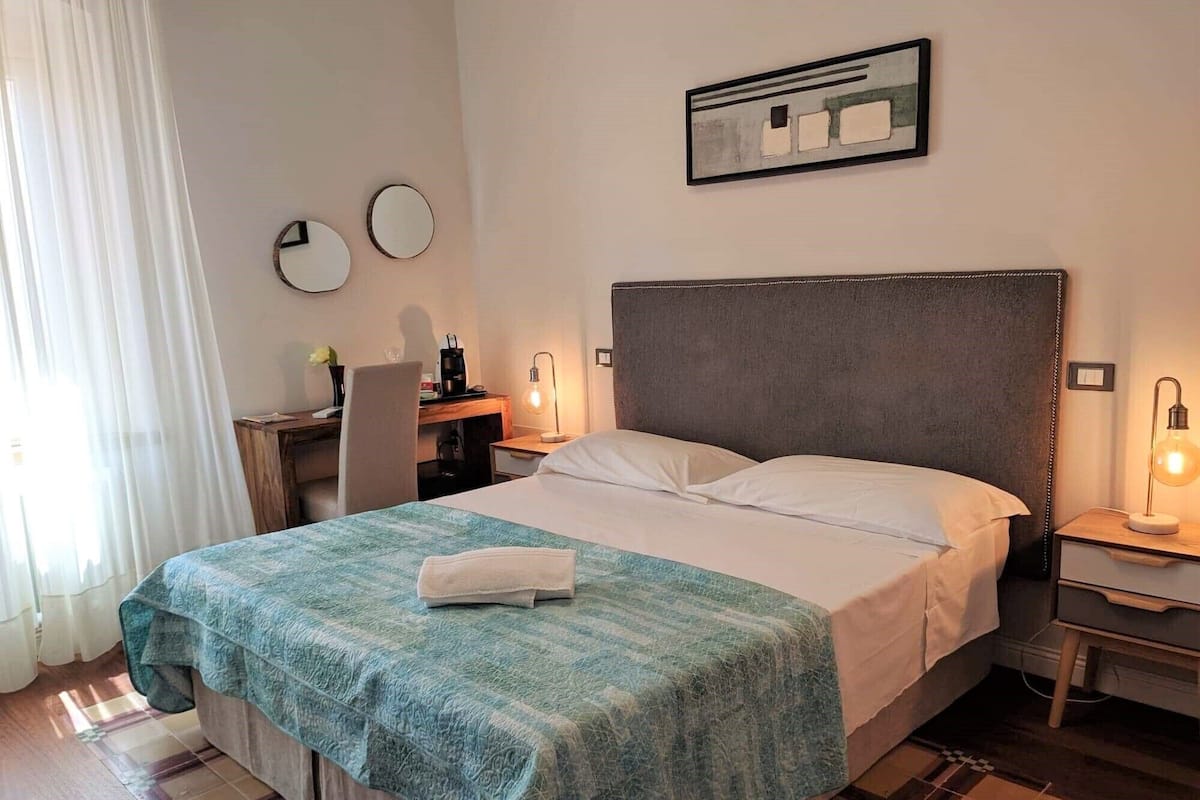 A hotel room with a bed, a desk, a chair, a nightstand, and a wall-mounted artwork.
