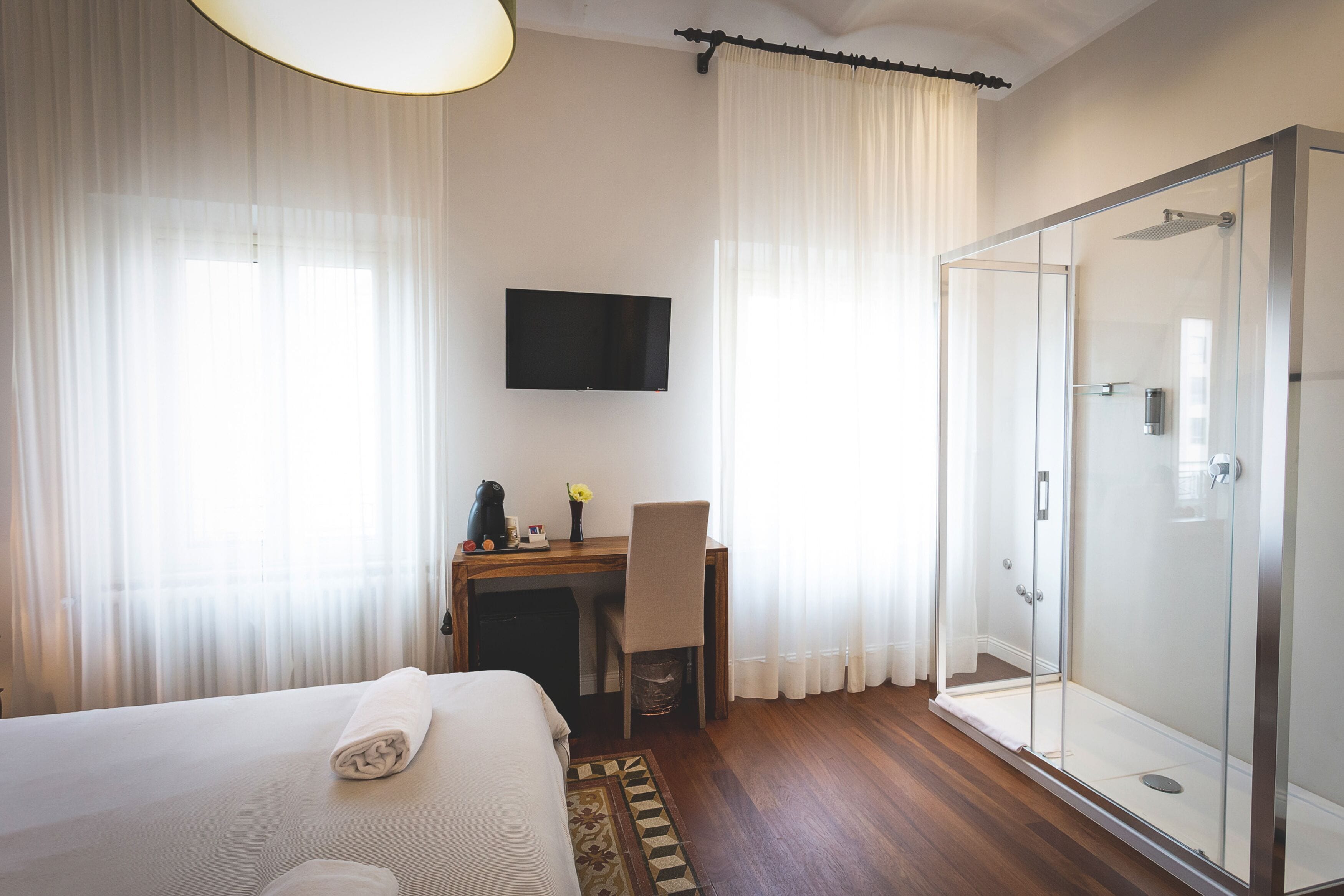 Een hotelkamer met een bed, een bureau met stoel, een televisie en een douche met glazen wand.