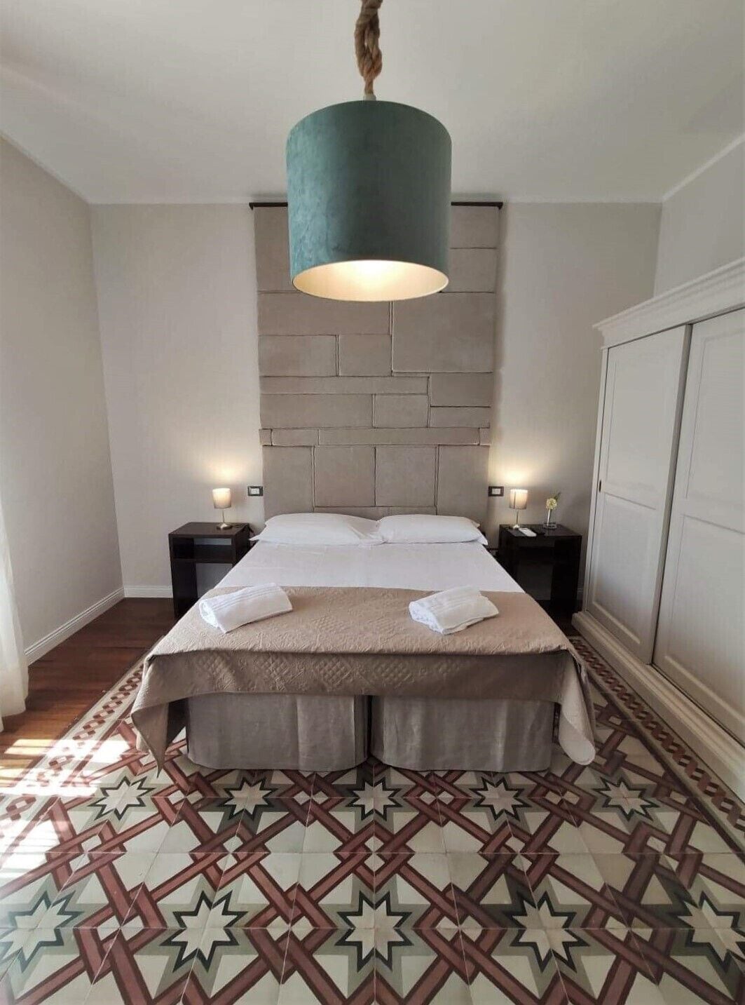 A bedroom with a bed, nightstands, a hanging light fixture, and patterned flooring.