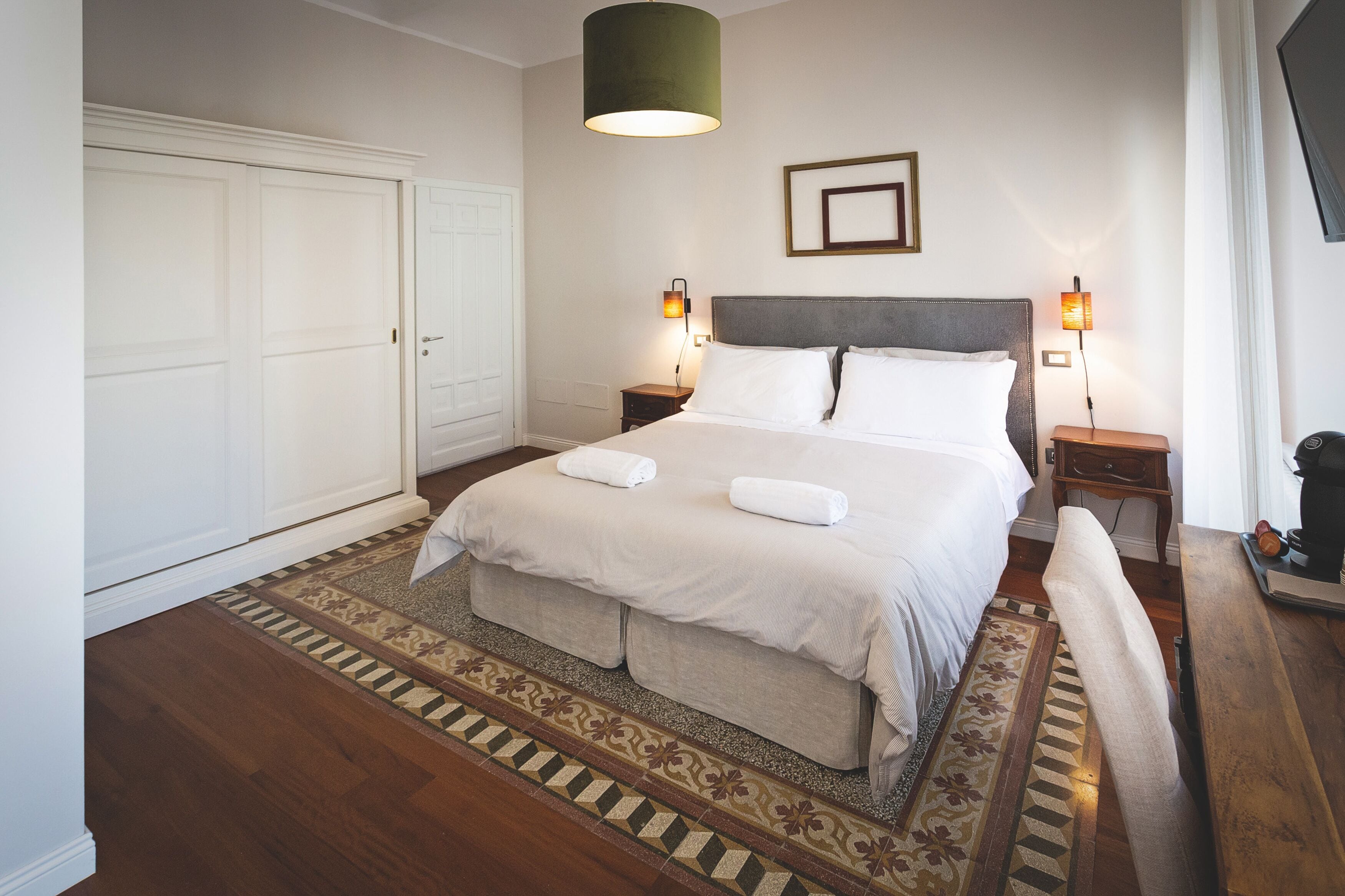 A neatly arranged bedroom with a large bed, two bedside tables with lamps, a wardrobe, and a patterned rug.