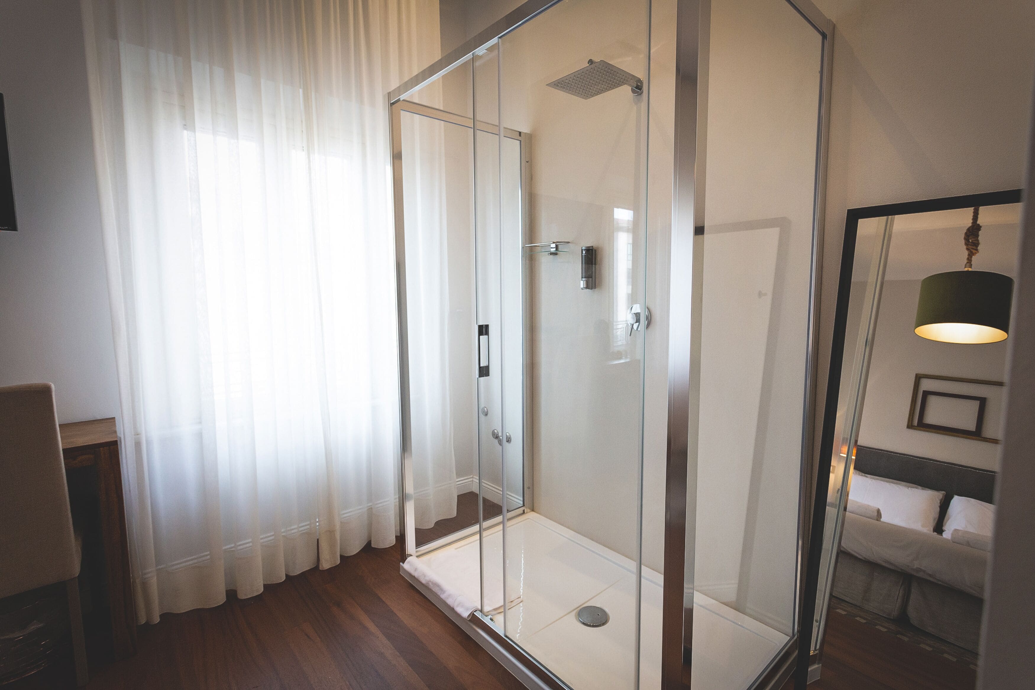 A modern bathroom with a glass shower enclosure, a wooden floor, and a bed with white bedding.