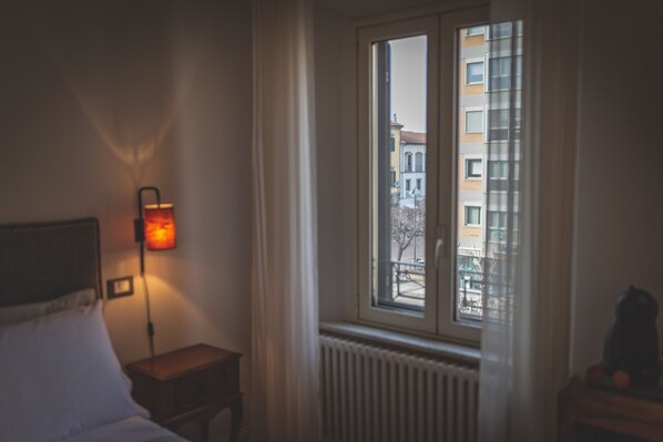 Habitación de hotel con cama, mesita de noche, lámpara, ventana con vista a edificios y radiador.