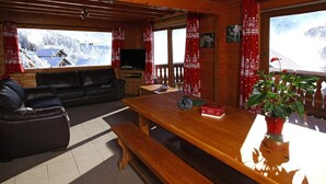 A cozy cabin interior with a wooden dining table, a leather sofa, and a large window with a view of snow-covered houses.