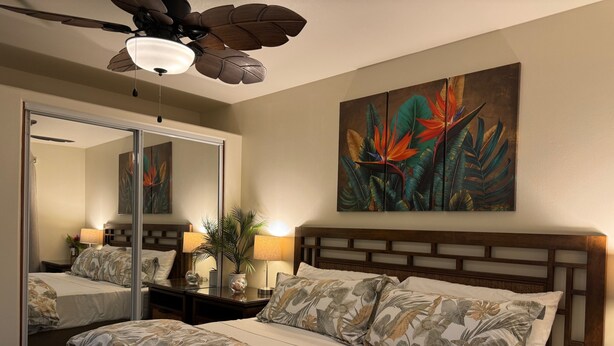 Quarto de hotel com uma cama grande, um ventilador de teto, um espelho e uma pintura com tema tropical na parede.