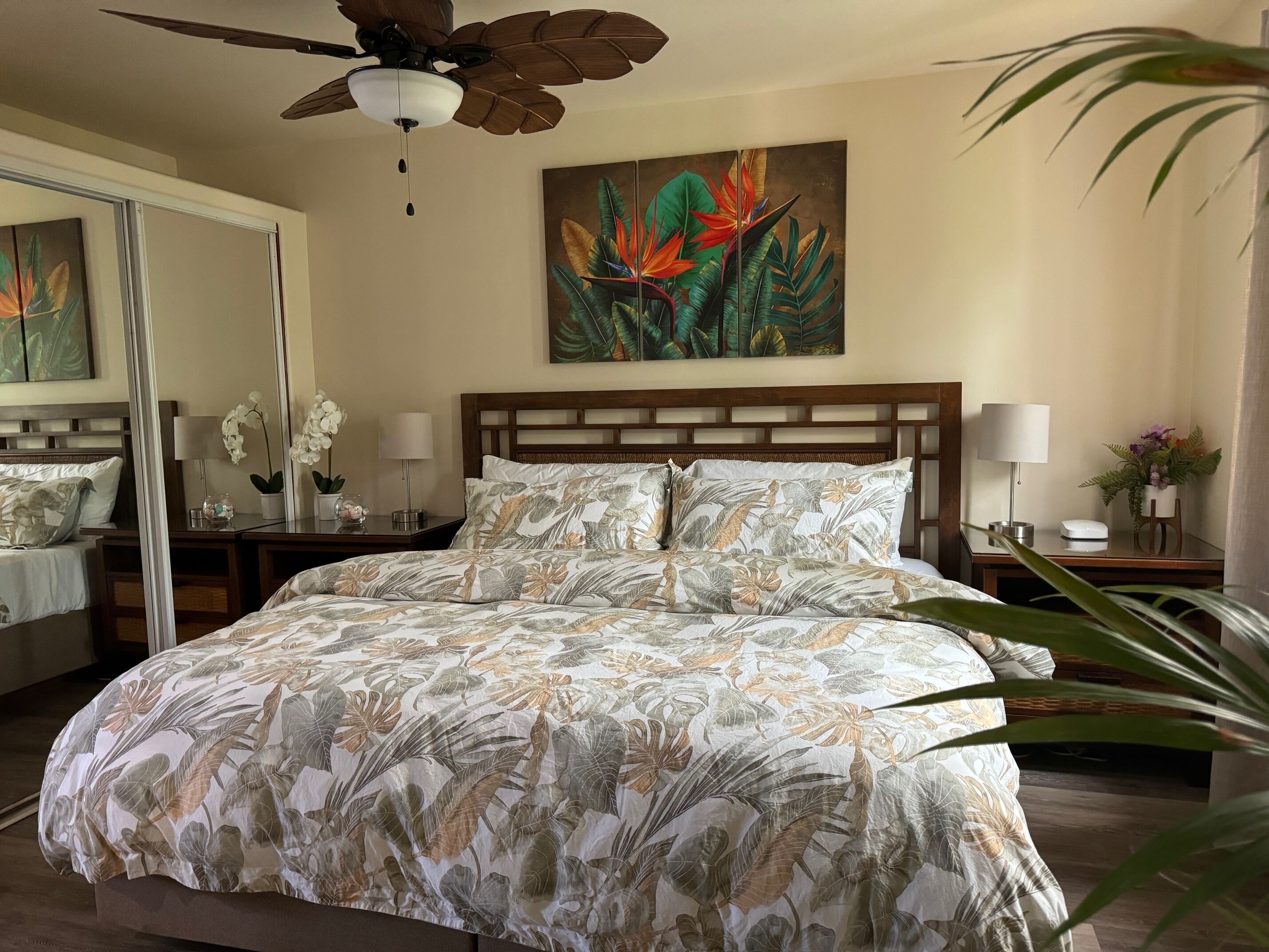 A hotel room with a large bed, a ceiling fan, a painting of tropical leaves, a nightstand with a lamp, and a mirror.