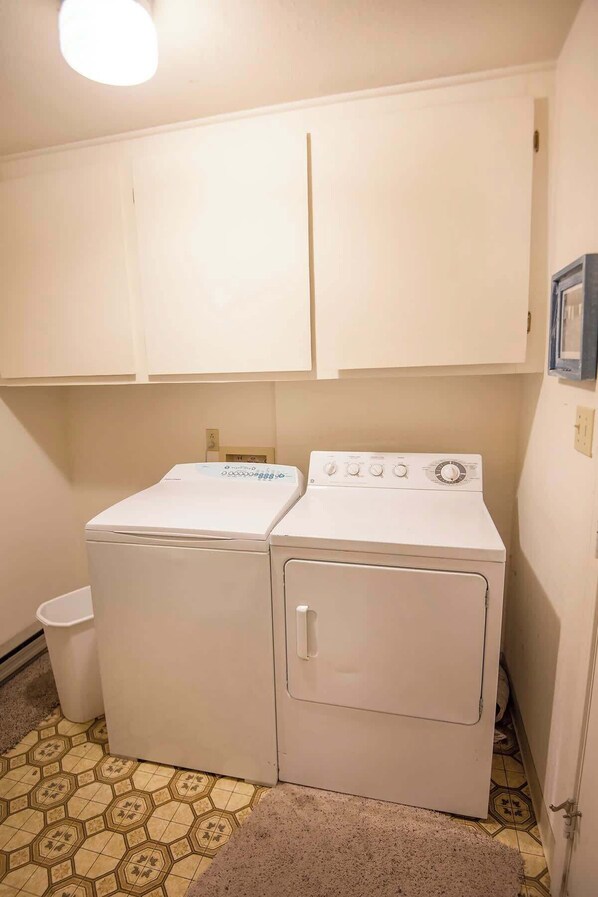 A laundry room with two white front-loading washing machines, a white trash bin, and a patterned floor. - Epic McCall Group Cabin | Close to Park, Lake,Town (McCall)
