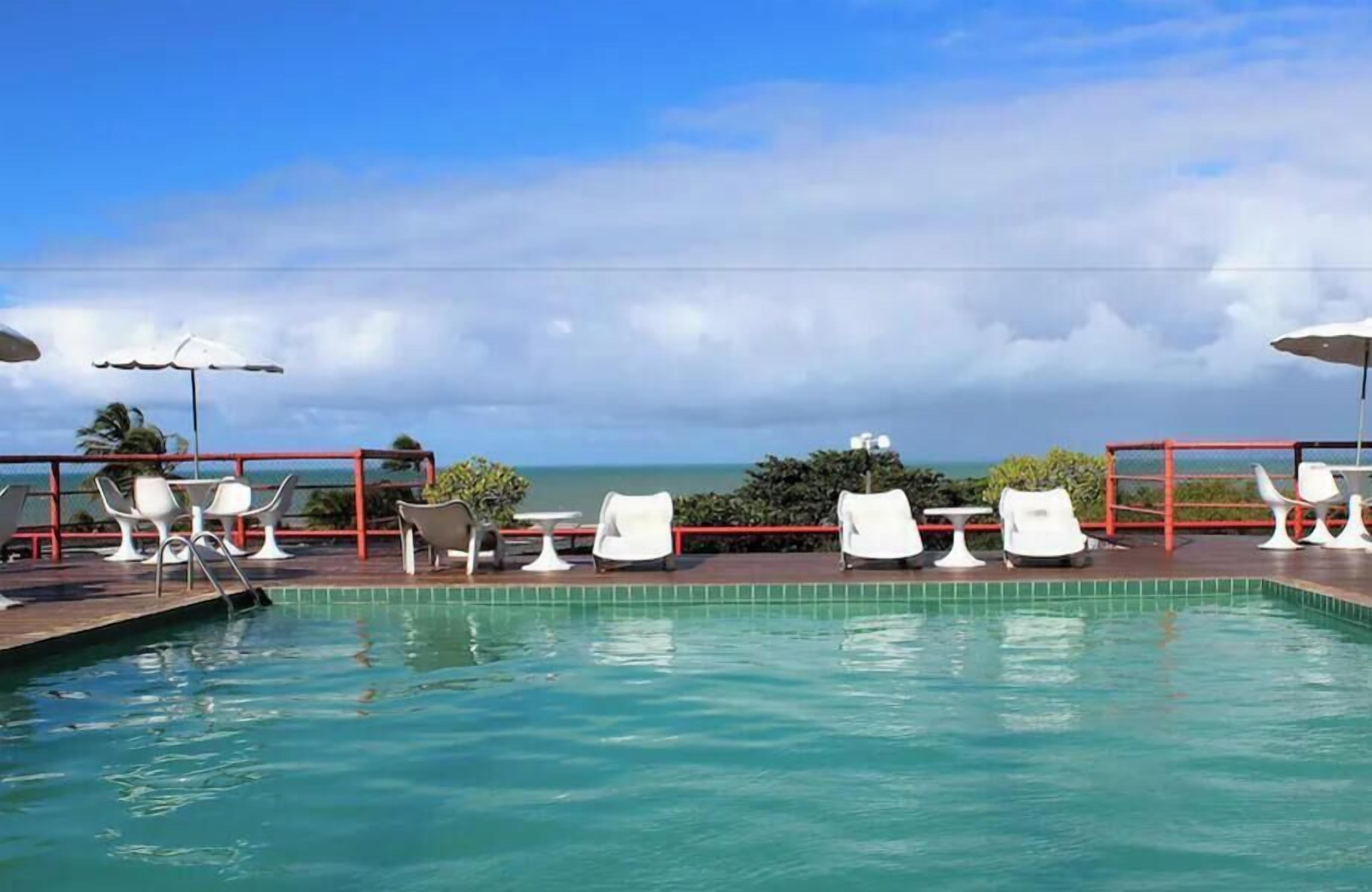 Une piscine bordée de chaises longues blanches et d’ombrelles, avec vue sur l’océan, et une rambarde rouge.
