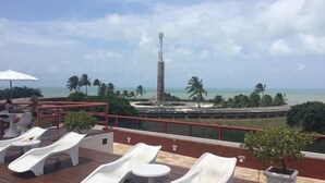 Une terrasse sur le toit, dotée de chaises longues, offrant une vue sur l’océan, des palmiers et une haute tour de communication.