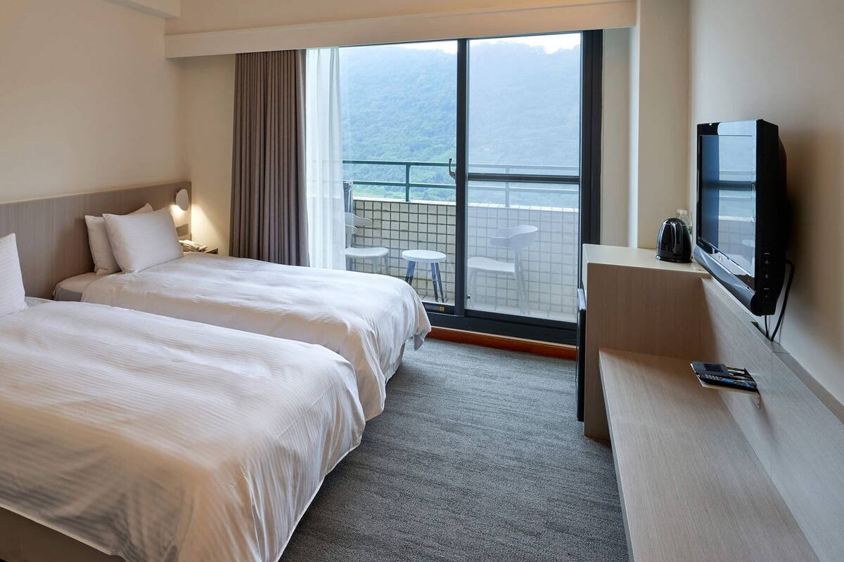 Ein Hotelzimmer mit zwei Betten, einem Fernseher, einem Schreibtisch und einem Balkon mit Aussicht.