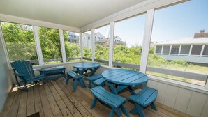 A covered patio with wooden flooring, blue outdoor furniture, and large windows offering a view of greenery and buildings. - Saltaire Cottage 11: 3BR Steps to Private Beach Access Screened Porch Pool (Kitty Hawk)