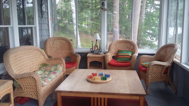 A cozy room with wicker furniture, a wooden table, and a view of trees through large windows. - Waterfront cottage on Lake Christopher, screened in porch w/ lake views, firepit (WOODSTOCK)