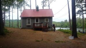 A cabin with a red roof, a deck, and a view of a lake surrounded by trees. - Waterfront cottage on Lake Christopher, screened in porch w/ lake views, firepit (WOODSTOCK)