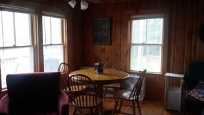 A wooden cabin interior with a round dining table surrounded by chairs, a ceiling fan, and large windows offering a view of the outdoors. - Waterfront cottage on Lake Christopher, screened in porch w/ lake views, firepit (WOODSTOCK)