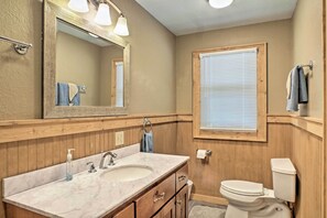 A bathroom with wooden paneling, a large mirror, a sink with a soap dispenser, a toilet, and a window with blinds. - The Aviator at Kudzu Cove Cabins Resort (Guntersville)