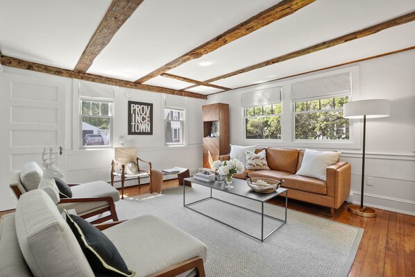 A spacious living room with a high ceiling, exposed wooden beams, a large window, and a cozy seating area with a sofa and chairs. - A Modern Antique Home - steps to Dock Square and harbor (Kennebunkport)