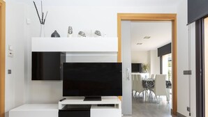 Uma sala de estar moderna com uma televisão de tela plana grande sobre um móvel branco, uma área de jantar com mesa e cadeiras, e vista para o cômodo adjacente.