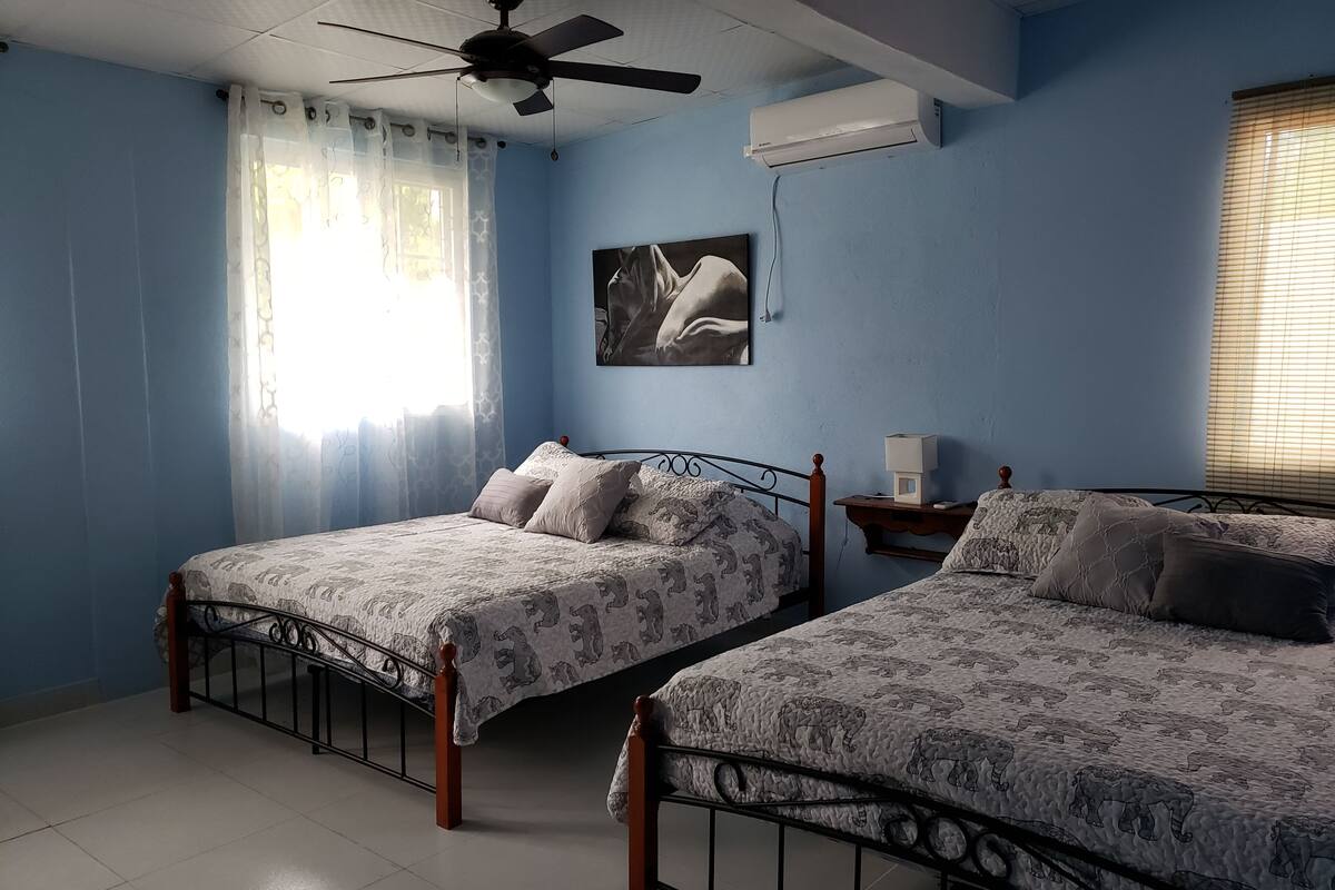 Un dormitorio con dos camas, un ventilador de techo, aire acondicionado y una ventana con cortinas.
