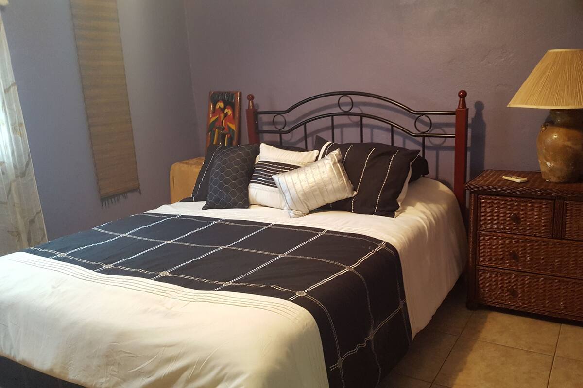Una cama bien hecha con colcha blanco y negro, una lámpara de noche y una mesita de noche de mimbre con lámpara.