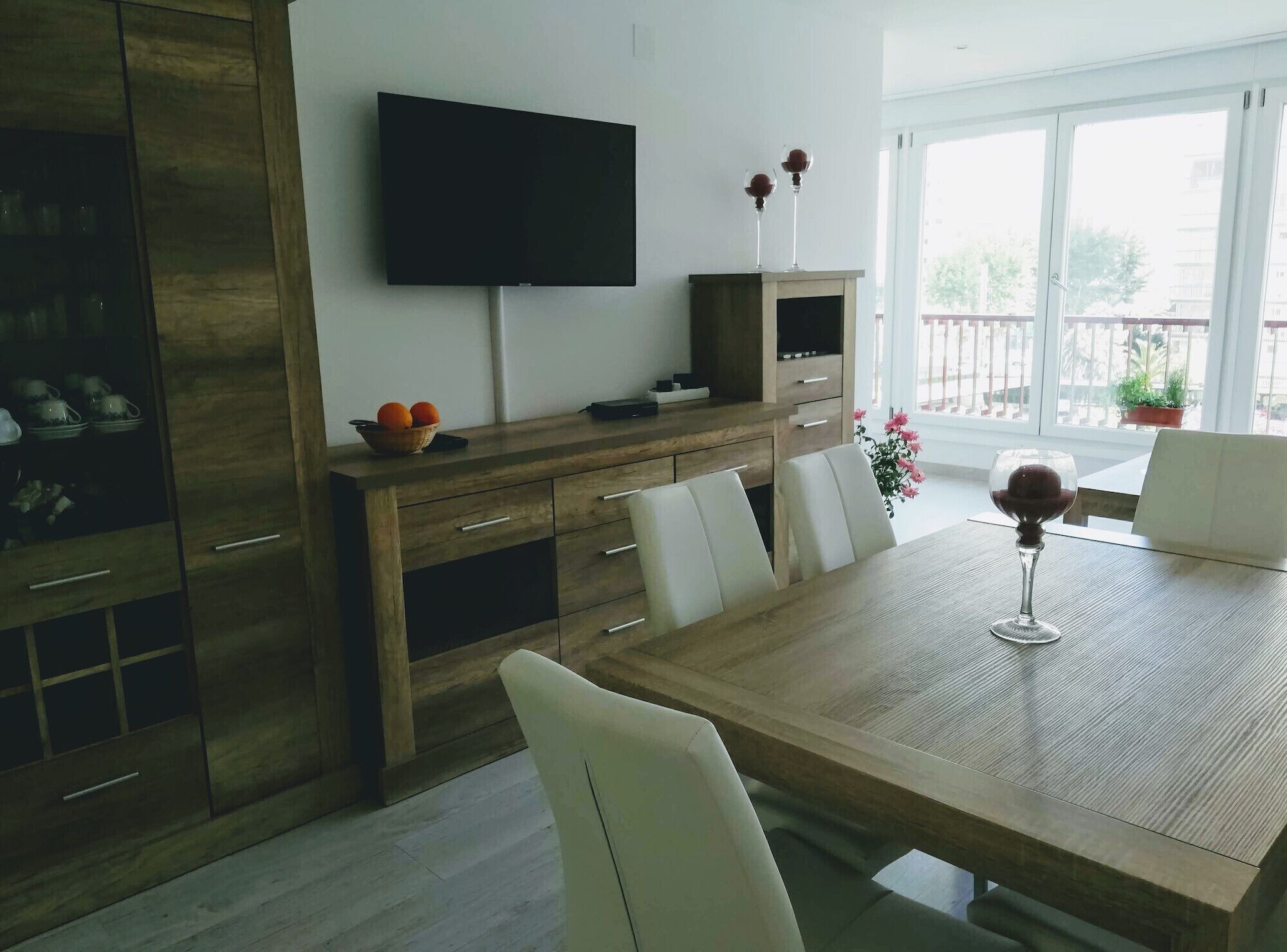 A modern dining area with a wooden table, white chairs, a flat-screen TV mounted on the wall, and a balcony with potted plants.