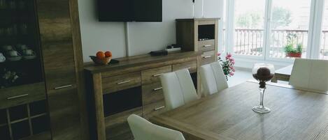 A modern dining area with a wooden table, white chairs, a flat-screen TV mounted on the wall, and a balcony with potted plants.