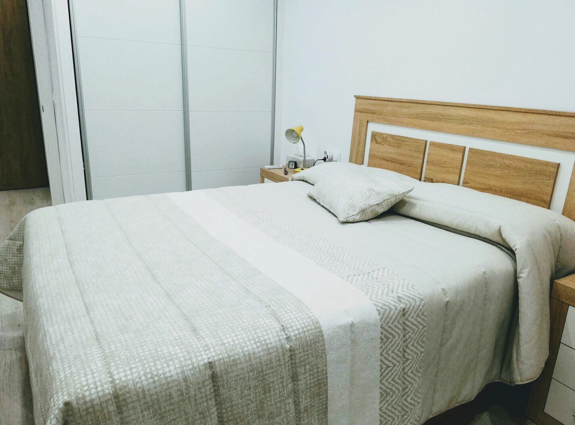 A neatly made bed with a wooden headboard, a bedside table with a lamp, and a white wall.