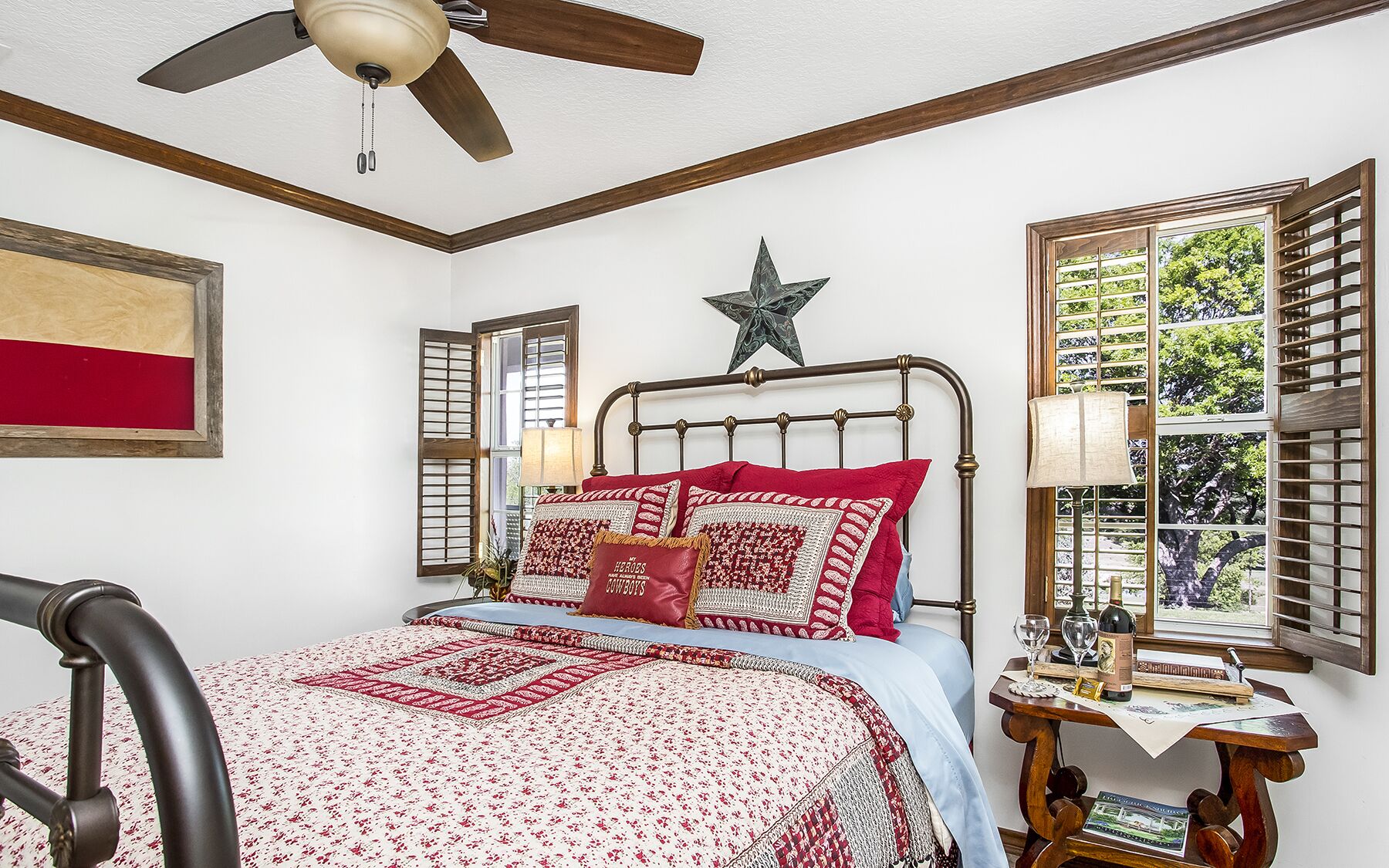 A bedroom with a bed, a ceiling fan, a star decoration, a window with shutters, and a bedside table with a lamp and a bottle.