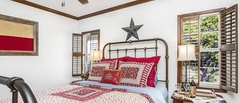 A bedroom with a bed, a ceiling fan, a star decoration, a window with shutters, and a bedside table with a lamp and a bottle.