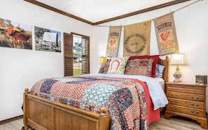 Une chambre avec un lit en bois, une table de chevet, une lampe et une commode. Les murs sont décorés, notamment avec une banderole et une affiche.
