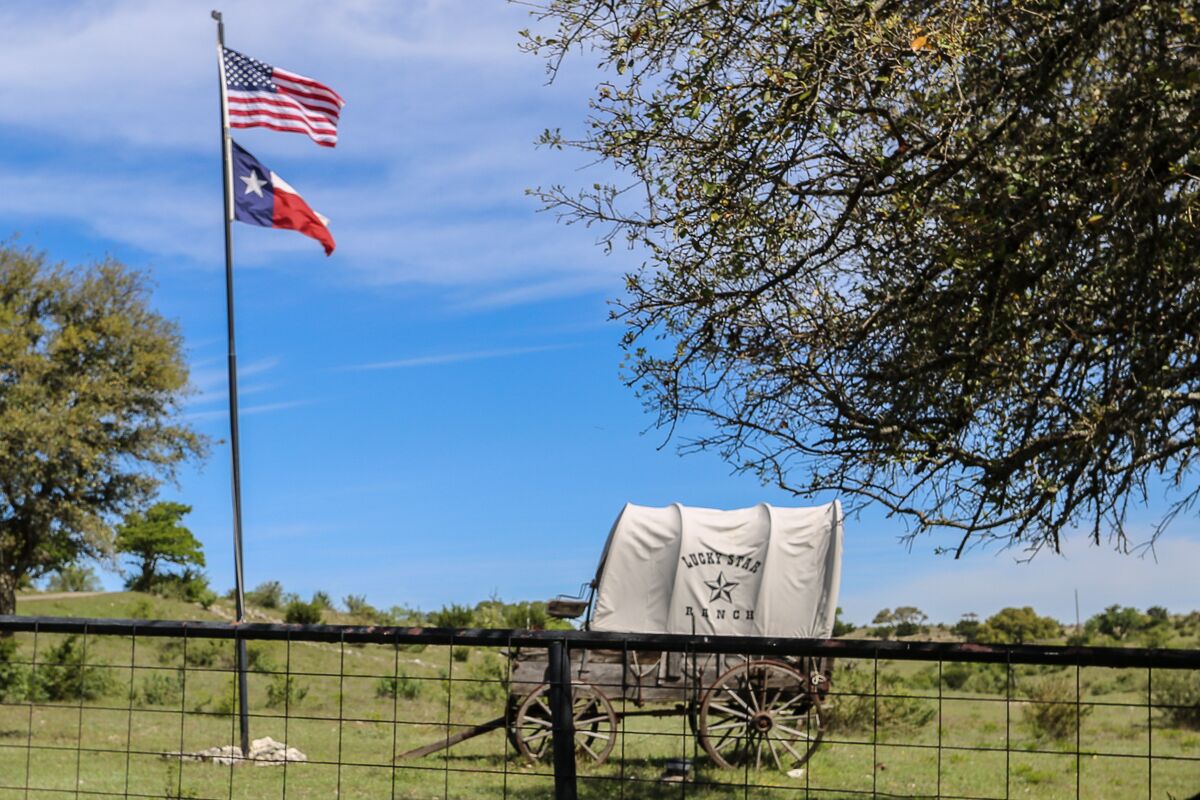 Un carro cobert amb la frase "Lucky Star Ranch", estacionat en una zona tancada per una tanca, amb una bandera dels Estats Units i una bandera de Texas al fons.