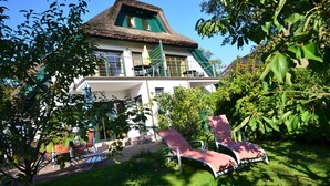 A large house with a thatched roof, a balcony, and a garden with lounge chairs.