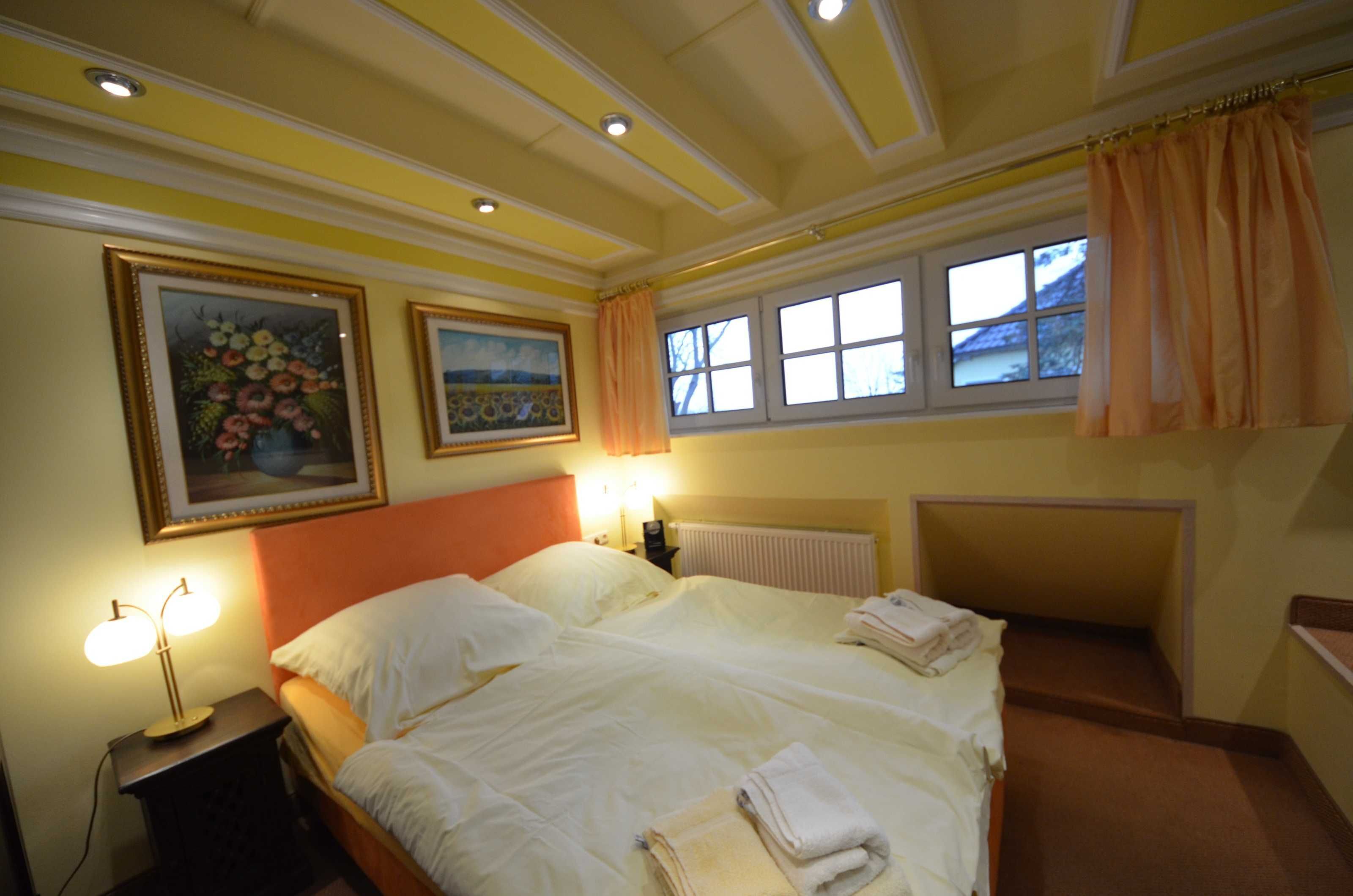 A double bed with white linens, a nightstand with a lamp, two framed floral paintings, a window with curtains, and a radiator.