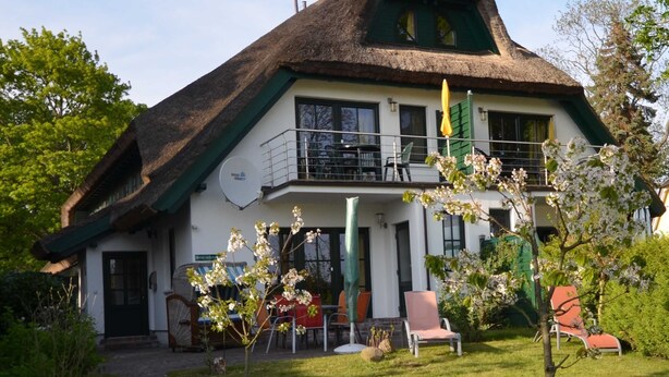 A two-story house with a thatched roof, a balcony with outdoor seating, and a garden with flowering plants.