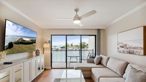 A modern living room with a large TV, a ceiling fan, a sofa, a coffee table, and a view of the ocean through the sliding glass doors. - Land's End 204 building 4 Premier|Large Pool|Steps to beach access! (Treasure Island)