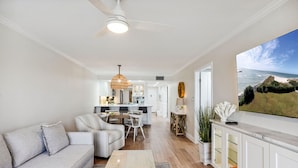 A modern living room with a sofa, coffee table, and a large TV screen displaying a scenic view. - Land's End 204 building 4 Premier|Large Pool|Steps to beach access! (Treasure Island)