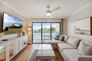 A modern living room with a large TV, a ceiling fan, a sofa, a coffee table, and a view of the ocean through the sliding glass doors. - Land's End 204 building 4 Premier|Large Pool|Steps to beach access! (Treasure Island)