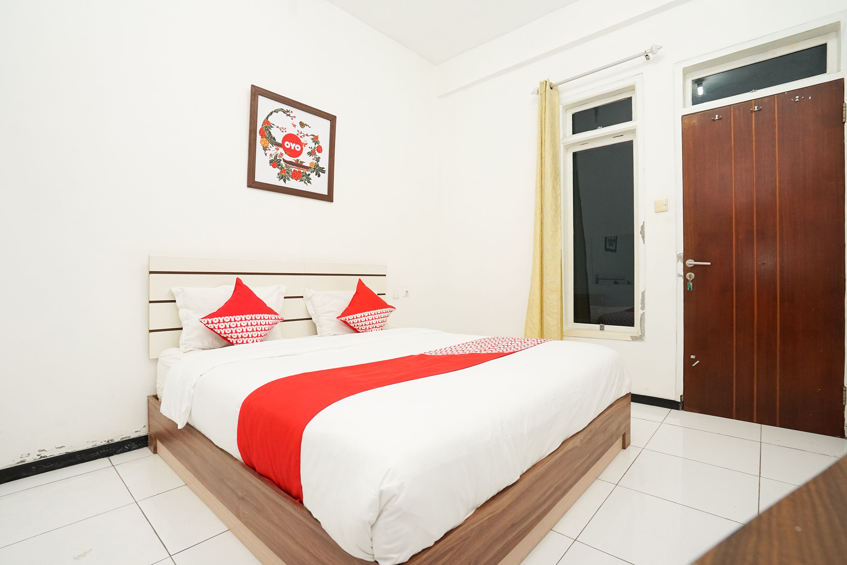 A bedroom with a bed, a wooden headboard, a red and white bedspread, a framed picture on the wall, and a wooden door.