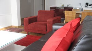 A living room with a red armchair, a grey sofa with red cushions, a glass coffee table, and a red rug.