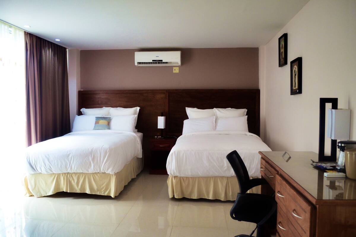 A hotel room with two beds, a desk, a chair, a dresser, and an air conditioning unit.