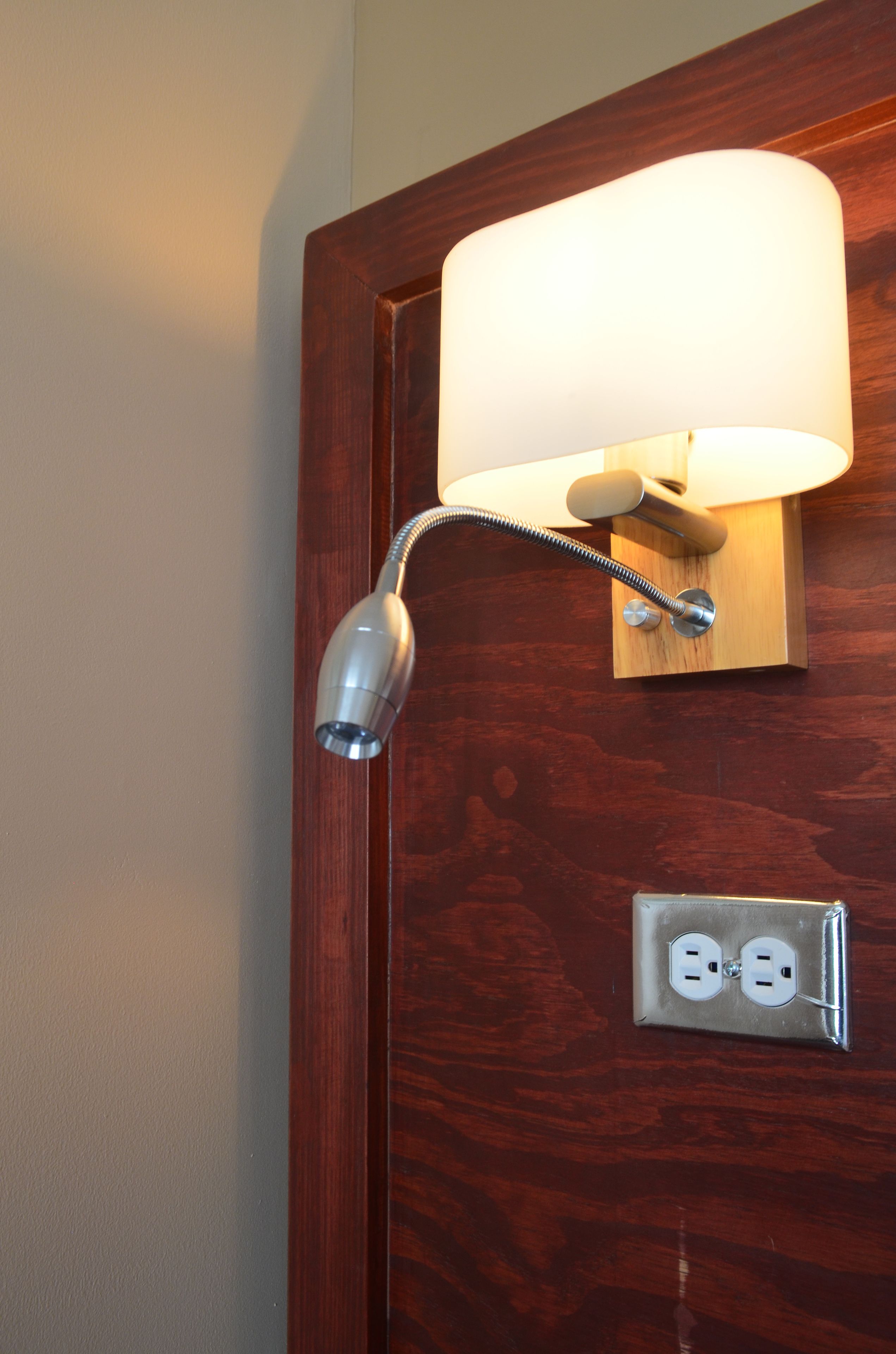 A wall-mounted lamp with a cylindrical shade and a flexible neck, attached to a wooden door with a visible electrical outlet below.