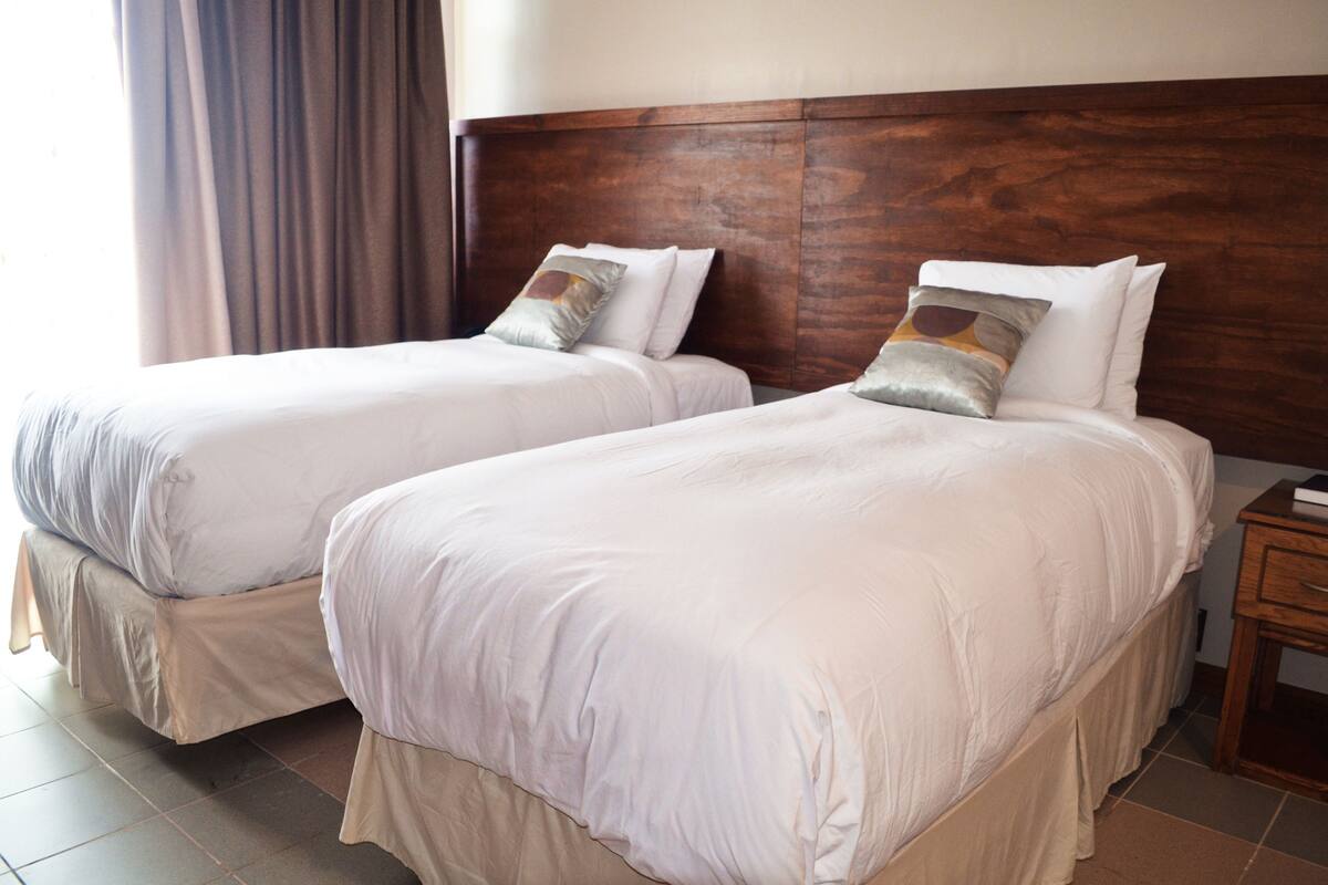 Two single beds with wooden headboards, a bedside table, and a window with curtains.