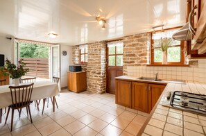 A kitchen with a stone wall, wooden cabinets, a tiled backsplash, a stove, and a dining area with a table and chairs. - Homerez - House in Beuzec-Cap-Sizun (Beuzec-Cap-Sizun)