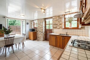 A kitchen with stone walls, wooden cabinets, a tiled floor, and a dining area with chairs and a table. - Homerez - House in Beuzec-Cap-Sizun (Beuzec-Cap-Sizun)