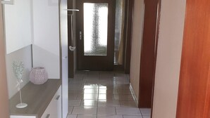 Um corredor com piso ladrilhado, uma geladeira branca, uma mesinha com um objeto decorativo e uma porta que dá para outro quarto.