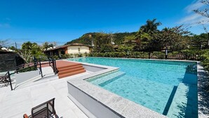 A clear swimming pool with a stone deck, steps leading into the pool, and a view of a hillside with trees and buildings in the background. - 2 bedrooms, 1 suite, 2 bathrooms, 100 m from the best location in Praia da Baleia. (São Sebastião)