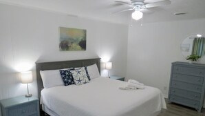 Una cama bien tendida con sábanas blancas, dos mesitas de noche con lámparas, un ventilador de techo y un aparador con espejo.